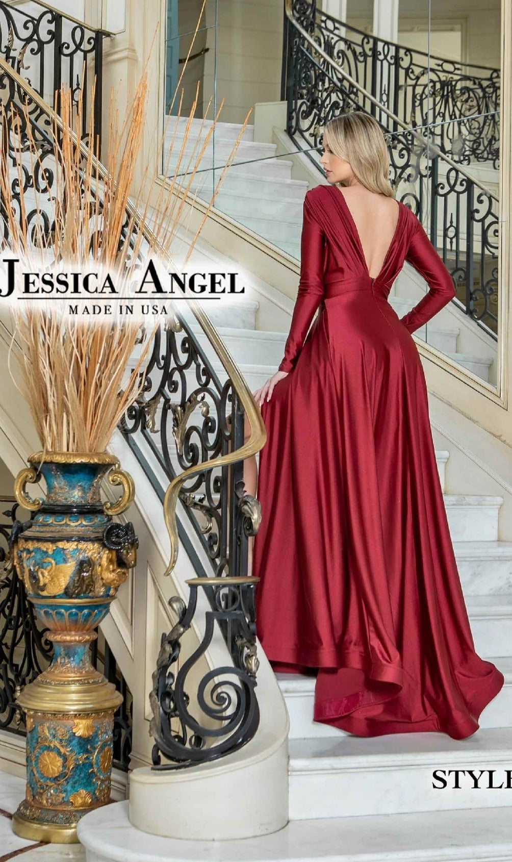 A woman in the Jessica Angel 796 Long Sleeve Long Formal Dress, deep red with an open back, stands on a staircase near an ornate vase and wrought iron railing.
